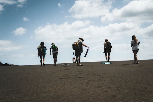 A group of people is walking up a sandy slope under a partly cloudy sky. Each person carries a backpack, and some have sandboards. The atmosphere is one of adventure and exploration.