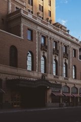 A historic brick building with ornate architectural details, featuring arched windows and decorative columns. The facade displays the inscription '1895 B.P.O.E No.6 1926' above the entrance. The building is bathed in warm sunlight with deep shadows, highlighting its vintage design.