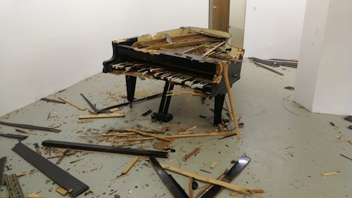 A grand piano is severely damaged and surrounded by scattered broken pieces of wood and debris in a room with plain white walls and a gray floor.