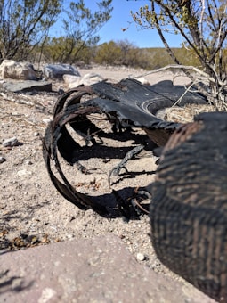 A worn and damaged tire lies on a dry, rocky landscape surrounded by sparse desert vegetation. The tire appears to be shredded and discarded amidst the arid terrain with a clear blue sky visible in the background.