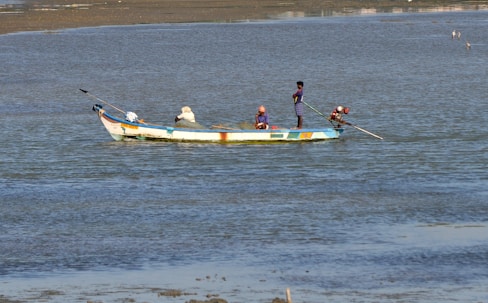 A small fishing boat is floating on calm waters. Three people are aboard, engaging in fishing activities. The boat is painted in bright colors, and the water shimmers under daylight.
