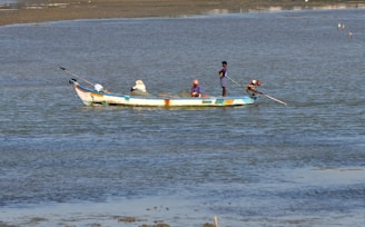 A small fishing boat is floating on calm waters. Three people are aboard, engaging in fishing activities. The boat is painted in bright colors, and the water shimmers under daylight.