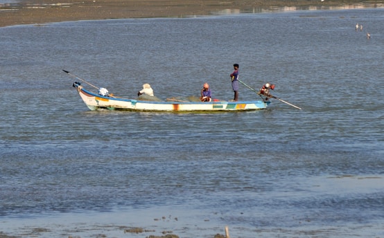 A small fishing boat is floating on calm waters. Three people are aboard, engaging in fishing activities. The boat is painted in bright colors, and the water shimmers under daylight.