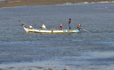 A small fishing boat is floating on calm waters. Three people are aboard, engaging in fishing activities. The boat is painted in bright colors, and the water shimmers under daylight.