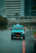 A blue taxi with a roof sign labeled 'Taksi' driving down a wet city street on a rainy day. The buildings in the background are modern, with glass and concrete facades, and other vehicles are visible further down the road. Reflections from the rain cover the road surface.
