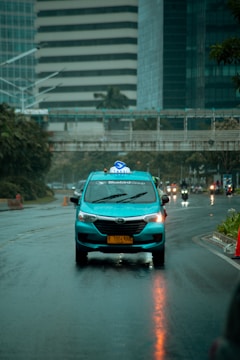 A blue taxi with a roof sign labeled 'Taksi' driving down a wet city street on a rainy day. The buildings in the background are modern, with glass and concrete facades, and other vehicles are visible further down the road. Reflections from the rain cover the road surface.