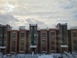 Modern three-story apartment buildings feature brick facades and tall windows. The structures have snowy rooftops and are set against a backdrop of cloud-filled skies, suggesting a cold or wintry atmosphere.