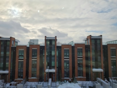 Modern three-story apartment buildings feature brick facades and tall windows. The structures have snowy rooftops and are set against a backdrop of cloud-filled skies, suggesting a cold or wintry atmosphere.