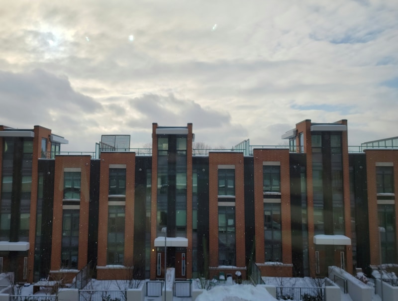 Modern three-story apartment buildings feature brick facades and tall windows. The structures have snowy rooftops and are set against a backdrop of cloud-filled skies, suggesting a cold or wintry atmosphere.