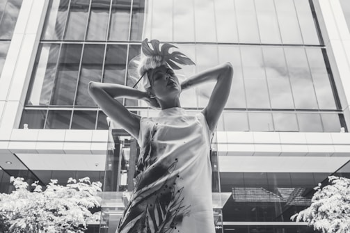 A person poses confidently in front of a modern glass building. The figure wears a stylish dress with expressive paint-like patterns and a decorative headpiece. The architectural features of the building are reflected on the glass surface, adding geometric elements to the backdrop.