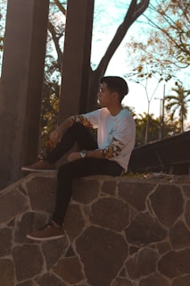 A person wearing a white shirt with patterned sleeves sits on a stone ledge. They appear to be deep in thought, looking towards the distance with trees and abstract structures in the background.