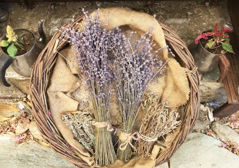 A wicker basket is filled with dried lavender, sage, and other herbs, laying on a burlap cloth. Two small metal pots with flowering plants flank the basket, resting on a stone surface adorned with scattered petals and leaves.