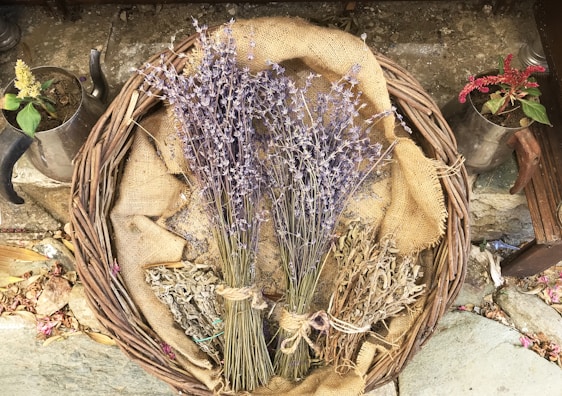 A wicker basket is filled with dried lavender, sage, and other herbs, laying on a burlap cloth. Two small metal pots with flowering plants flank the basket, resting on a stone surface adorned with scattered petals and leaves.