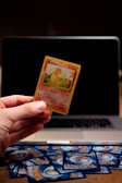 A hand is holding a Pokémon trading card featuring a character named Glumanda. The card is in the foreground with a laptop in the background. Several Pokémon card backs are scattered on a wooden surface.