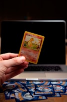 A hand is holding a Pokémon trading card featuring a character named Glumanda. The card is in the foreground with a laptop in the background. Several Pokémon card backs are scattered on a wooden surface.