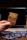 A hand is holding a Pokémon trading card featuring a character named Glumanda. The card is in the foreground with a laptop in the background. Several Pokémon card backs are scattered on a wooden surface.