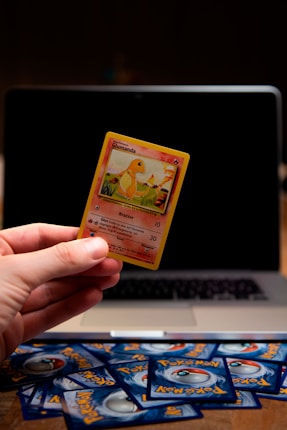 A hand is holding a Pok&eacute;mon trading card featuring a character named Glumanda. The card is in the foreground with a laptop in the background. Several Pok&eacute;mon card backs are scattered on a wooden surface.