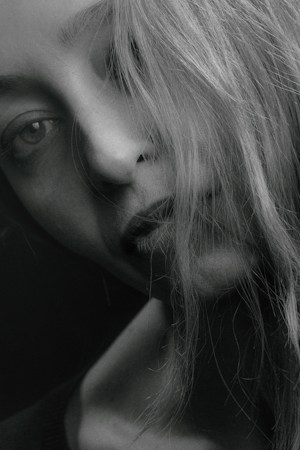 A close-up black and white portrait of a person's face partially obscured by strands of light-colored hair. The person's expression appears somber or thoughtful. The skin tones and hair textures are captured in fine detail.