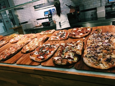 A variety of rectangular pizzas displayed on wooden boards in a pizzeria setting. Each pizza is topped with different ingredients like vegetables, meats, and cheeses. A person in a white shirt is visible in the background, likely preparing more food or handling customer orders at the counter.