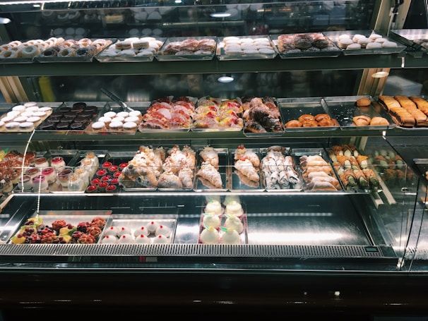 A well-lit pastry display case featuring an array of baked goods. The assortment includes croissants, cannoli, tarts, and various pastries decorated with powdered sugar, fruit, and chocolate. The shelves are organized with different kinds of sweets, creating an inviting and appetizing presentation.