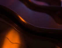 Curved, abstract shapes with smooth textures are bathed in warm light, creating a mesmerizing interplay of shadows and highlights. The surface appears glossy, reflecting light softly, with a gentle blend of colors creating a soothing and intriguing visual effect.