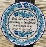 A round ceramic tile features blue and green floral patterns encircling a Spanish inscription. The script is handwritten in black ink and contrasts with the white background of the plate. The intricate design demonstrates traditional craftsmanship.