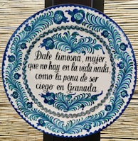 A round ceramic tile features blue and green floral patterns encircling a Spanish inscription. The script is handwritten in black ink and contrasts with the white background of the plate. The intricate design demonstrates traditional craftsmanship.