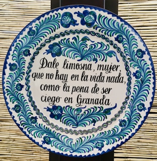 A round ceramic tile features blue and green floral patterns encircling a Spanish inscription. The script is handwritten in black ink and contrasts with the white background of the plate. The intricate design demonstrates traditional craftsmanship.