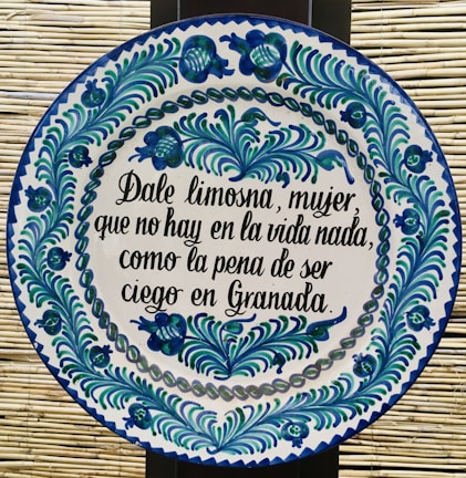 A round ceramic tile features blue and green floral patterns encircling a Spanish inscription. The script is handwritten in black ink and contrasts with the white background of the plate. The intricate design demonstrates traditional craftsmanship.