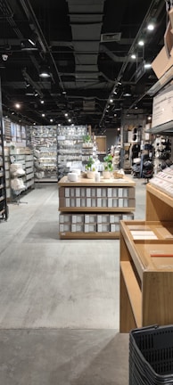 A well-organized interior of a retail store featuring neatly arranged shelves filled with various home goods and products. The aisle is wide, with wooden display tables showcasing white containers and decorative green plants. Ceiling lights provide ample illumination, and the store has a contemporary, clean design.