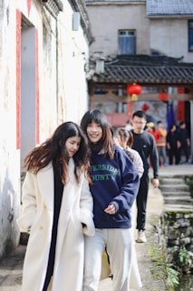 A group of people walk along a narrow outdoor path next to a stone building with traditional architecture. The two women in the front are smiling and walking closely together. The background features red lanterns and several other people in casual clothing.