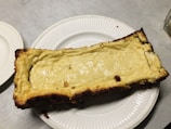 A rectangular cheesecake with a golden brown crust edge, placed on a white plate. The cheesecake has a smooth, glossy surface with a light yellow color. It rests on a light gray table surface.