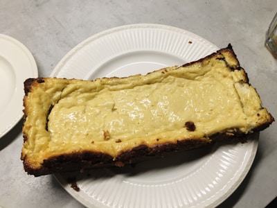 A rectangular cheesecake with a golden brown crust edge, placed on a white plate. The cheesecake has a smooth, glossy surface with a light yellow color. It rests on a light gray table surface.