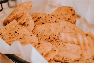 A close-up view of seasoned, crispy potato chips in a basket lined with white paper. The chips have a golden-brown color with visible specks of herbs and seasoning, suggesting a savory flavor.