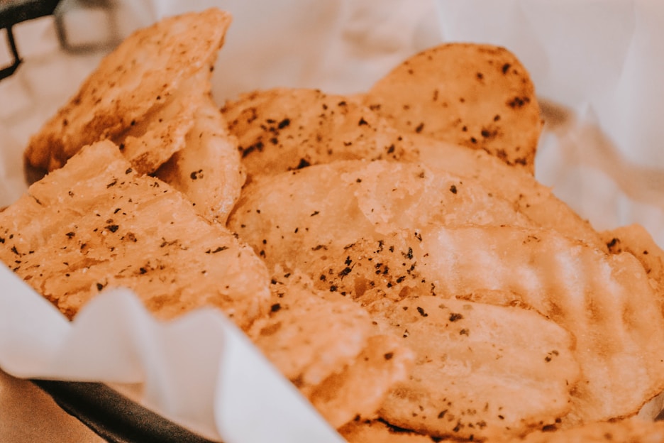 A close-up view of seasoned, crispy potato chips in a basket lined with white paper. The chips have a golden-brown color with visible specks of herbs and seasoning, suggesting a savory flavor.