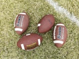 Four American footballs are placed on a grassy field with one of them lying near a white painted line. The footballs are brown with white stripes and feature the branding 'Franklin GRIP-RITE' on their surface.