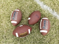 Four American footballs are placed on a grassy field with one of them lying near a white painted line. The footballs are brown with white stripes and feature the branding 'Franklin GRIP-RITE' on their surface.