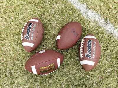 Four American footballs are placed on a grassy field with one of them lying near a white painted line. The footballs are brown with white stripes and feature the branding 'Franklin GRIP-RITE' on their surface.