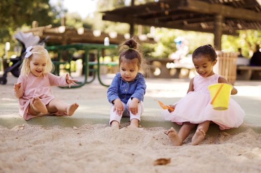 Three young children are sitting and playing in a sandbox in a park. One child with blonde hair and a pink dress is reaching out towards the sand. The middle child, wearing a denim jacket and white pants, is looking intently at the sand. The third child, in a pink dress, is holding a yellow bucket and an orange spade, smiling as she digs in the sand. The background features a green picnic table and lush greenery with a sunny atmosphere.