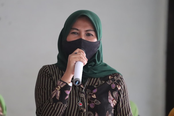 A person wearing a green headscarf and a black face mask holds a microphone. The individual is dressed in a patterned outfit featuring various designs and appears to be speaking or presenting something.