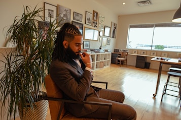 A man in a suit sits thoughtfully in a modern, stylish office space. The room features wooden flooring, a variety of framed artworks on the wall, and a large potted plant. There's a window providing natural light, and a long desk with chairs and some office items.