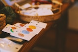 Colorful enamel pins are arranged on display cards, placed on a wooden surface. Behind them, there is a woven basket containing various other items partially blurred in the background.