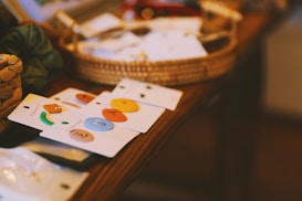 Colorful enamel pins are arranged on display cards, placed on a wooden surface. Behind them, there is a woven basket containing various other items partially blurred in the background.