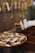 A collection of gold jewelry displayed on a wooden dish with price tags attached. Blurred background shows more hanging jewelry pieces against a wall.