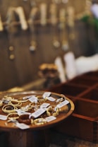 A collection of gold jewelry displayed on a wooden dish with price tags attached. Blurred background shows more hanging jewelry pieces against a wall.