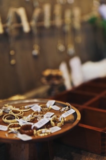 A collection of gold jewelry displayed on a wooden dish with price tags attached. Blurred background shows more hanging jewelry pieces against a wall.