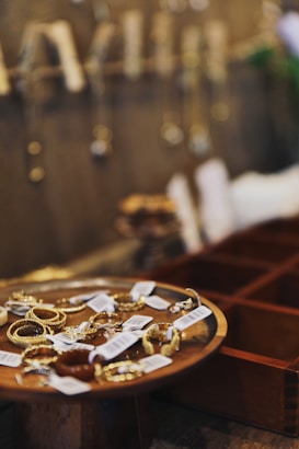 A collection of gold jewelry displayed on a wooden dish with price tags attached. Blurred background shows more hanging jewelry pieces against a wall.