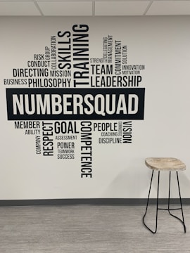 A wall of typography art featuring motivational words and phrases such as 'Skills', 'Training', 'Leadership', and 'Team'. The word 'NUMBERSQUAD' is prominently displayed in the center. Adjacent to the wall is a tall stool with a wooden seat and metal legs on a light, laminate floor.