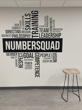 A wall of typography art featuring motivational words and phrases such as 'Skills', 'Training', 'Leadership', and 'Team'. The word 'NUMBERSQUAD' is prominently displayed in the center. Adjacent to the wall is a tall stool with a wooden seat and metal legs on a light, laminate floor.