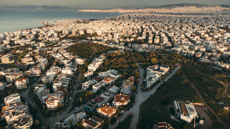 The southern suburbs of Athens from the air, white low-rise and pine running down to the Saronic Gulf.